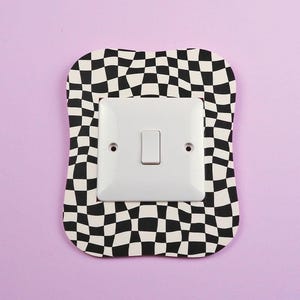 Può includere: Un interruttore bianco è montato su una placca decorativa a scacchi bianchi e neri di forma irregolare. Lo sfondo è di un viola chiaro uniforme. La placca è un oggetto decorativo.
