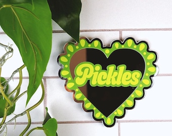May include: A heart-shaped mirror with a green and yellow frame. The mirror has the word "Pickles" written in yellow letters on the reflective surface.