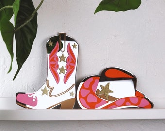 May include: A pink and white cowboy boot and a pink and red cowboy hat with gold stars. The boot and hat are made of mirrored acrylic.