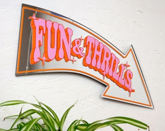 May include: A silver mirrored arrow sign with the text "FUN & THRILLS" in pink and orange letters. The sign is mounted on a white wall.