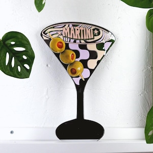 Può includere: Un bicchiere da martini bianco e nero con un motivo a spirale rosa e bianco. Il bicchiere ha tre olive verdi su uno stuzzicadenti. Sul bicchiere è scritta la parola "MARTINI".