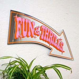 May include: A silver mirrored arrow sign with the text "FUN & THRILLS" in pink and orange letters. The sign is mounted on a white wall.