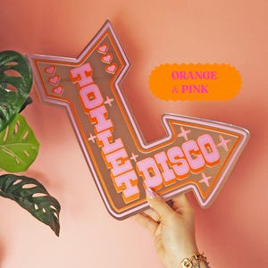 Puede incluir: Un letrero en forma de flecha con las palabras "HOHHH DISCO" en rosa y naranja. El letrero tiene un fondo marrón y está decorado con corazones y estrellas. El letrero se sostiene contra una pared rosa.