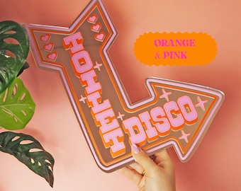May include: An arrow-shaped sign with the words "HOHHH DISCO" in pink and orange. The sign has a brown background and is decorated with hearts and stars. The sign is held against a pink wall.