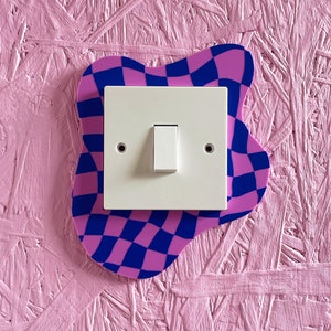 May include: A white light switch with a toggle button is mounted on a pink and blue checkered sticker. The sticker is shaped like a cloud and is attached to a pink wall.