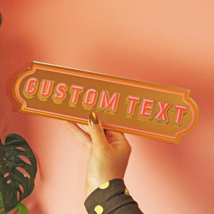May include: A gold-colored sign with the words "CUSTOM TEXT" in pink and orange lettering. The sign has a decorative border and is held up against a pink wall. Green leaves are visible in the background.