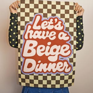 Può includere: Un canovaccio da cucina a scacchi beige e marrone con il testo "Let's have a Beige Dinner" in un font retrò. L'asciugamano è tenuto da una persona che indossa una gonna di jeans e una camicia nera a pois gialli.