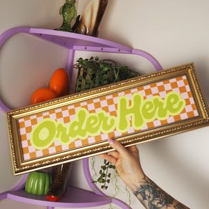 Op de afbeelding: Een goudomrand bord met de woorden "Order Here" in limoengroen, tegen een roze en oranje geruite achtergrond. Het bord wordt voor een paars rek met decoratieve items gehouden.