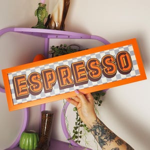 May include: An orange framed sign with the word "ESPRESSO" in brown and orange retro font. The background is a checkered pattern of gray and white. The sign is being held up in front of a purple shelf with decorative items.