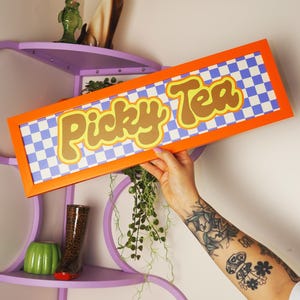 Può includere: Un cartello rettangolare arancione con la scritta "Picky Tea" in un font retrò su sfondo a scacchi blu e bianco. Il cartello è tenuto davanti a una mensola viola con oggetti decorativi.