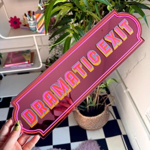 Puede incluir: Un letrero acrílico burdeos con las palabras "DRAMATIC EXIT" en letras 3D coloridas. El letrero tiene un borde rosa y naranja. Se muestra frente a un suelo a cuadros y una planta.