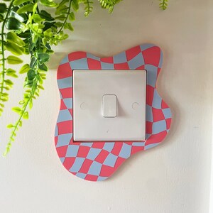 May include: A white light switch with a pink and blue checkered border. The border is a wavy, abstract shape.