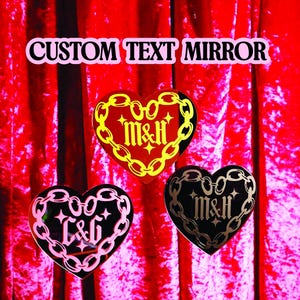 Puede incluir: Tres espejos en forma de corazón con bordes de cadena y texto personalizado. Los espejos son rojos, negros y rosas, con texto dorado y plateado. El texto "CUSTOM TEXT MIRROR" está en la parte superior.