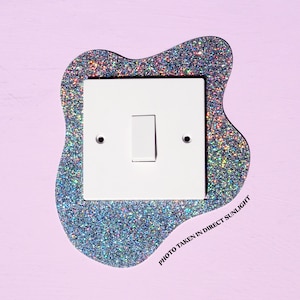 Puede incluir: Un interruptor de luz blanco con un borde de purpurina plateada. El borde tiene forma de nube. El texto "PHOTO TAKEN IN DIRECT SUNLIGHT" está impreso debajo del interruptor.