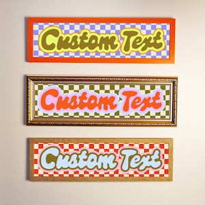 May include: Three framed wall art pieces with the words "Custom Text" in a retro font. Each piece has a different colour scheme and chequered background. The frames are orange, gold, and wood-toned.