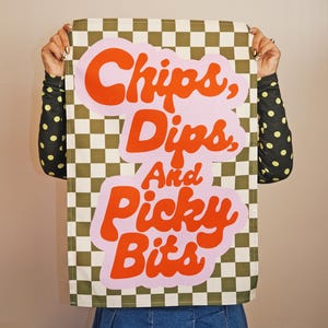 Könnte beinhalten: Ein rechteckiges Küchentuch mit einem karierten olivgrünen und weißen Muster. Das Tuch trägt den Text "Chips, Dips, And Picky Bits" in orangefarbenen Buchstaben mit rosa Umrandung. Das Tuch wird von einer Person gehalten.