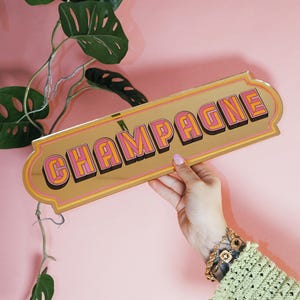Puede incluir: Un letrero dorado con la palabra "CHAMPAGNE" en letras rosas y naranjas. El letrero tiene un borde decorativo y se sostiene sobre un fondo rosa. Se ven hojas verdes en el fondo.