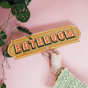 May include: A gold-colored metal bathroom sign with the word "BATHROOM" in orange letters with a green outline. The sign is held against a pink background, with green leafy plants in the upper left corner.