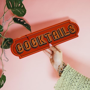 May include: A rectangular orange metal sign with the word "COCKTAILS" in black and orange lettering. The sign is held by a person's hand against a pink background, with green leafy plants in the upper left corner.