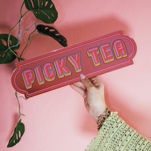 Può includere: Un'insegna rettangolare con le parole "PICKY TEA" in un font retrò. L'insegna è tenuta da una persona con una manica all'uncinetto verde e braccialetti dorati. Lo sfondo è di colore rosa pieno con una pianta verde.