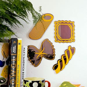 May include: A selection of pasta-shaped wall decorations, including rigatoni, farfalle, and fusilli. The decorations are yellow and brown, with a reflective surface. A mug with a floral design and a coaster are in the foreground.