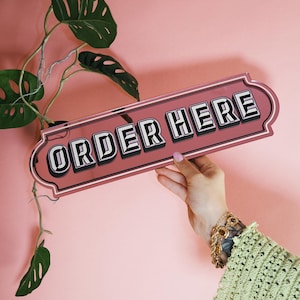 Op de afbeelding: Een roze rechthoekig bord met de tekst "ORDER HERE" in vette, zwart-witte letters. Het bord wordt vastgehouden door een hand met een gouden armband, tegen een roze achtergrond. Groene plantenbladeren zijn op de achtergrond te zien.