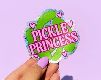 Könnte beinhalten: Ein Aufkleber mit dem Text "PICKLE PRINCESS" in Rosa, umgeben von grünen Gurkenillustrationen und rosa Herzen. Der Aufkleber hat einen holografischen, glitzernden Rand. Der Hintergrund ist ein helles Lila.