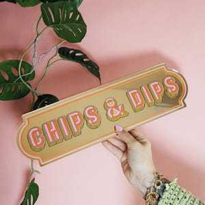 May include: A gold-colored sign with the words "CHIPS & DIPS" in pink and orange lettering. The sign has a decorative border and is held against a pink background. A green plant is in the background.