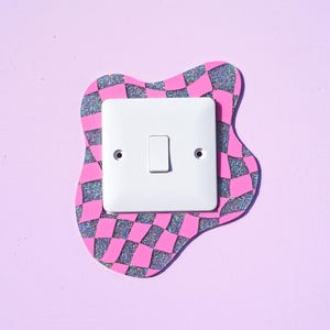 Può includere: Un interruttore della luce bianco con un interruttore a levetta è montato su una placca decorativa a scacchi rosa e neri con un bordo glitterato. La placca ha una forma irregolare e ondulata ed è posta su uno sfondo viola chiaro.