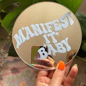 May include: A round, gold-colored mirror with the phrase "MANIFEST IT BABY" in white lettering. The lettering is in a retro, bubbly font with stars around the words.