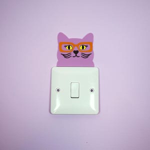 Puede incluir: Una cubierta de interruptor de luz caprichosa con la forma de una cara de gato púrpura con gafas naranjas. El gato está colocado encima de un interruptor de luz blanco en una pared lavanda. El gato tiene ojos amarillos y bigotes negros.