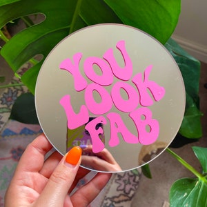 May include: A round mirror with a pink "You Look Fab" design. The mirror is held in a hand with orange nail polish.