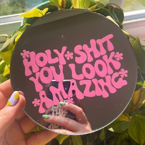 May include: A round mirror with a pink, cursive message that reads "*HOLY* *SHIT* YOU LOOK *AMAZING*" on a black background.