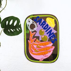 May include: A mirrored wall decor featuring a stylised illustration of a can of sardines. The can is brown with a green border and features a purple starburst, a lemon slice, two green olives, and a pull tab. The word "SARDINES" is written in blue letters on the can. Three pink sardines with orange fins are swimming inside the can.
