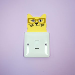 Puede incluir: Una cubierta de interruptor con forma de gato amarillo con bigotes y gafas negras está montada sobre un interruptor blanco en una pared lila. La cara del gato es el punto focal, agregando un toque lúdico a la decoración de la habitación.