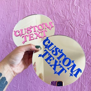 May include: Two round, gold-toned mirrors with the words "CUSTOM TEXT" in pink and blue, with star accents. The mirrors are held against a textured, light purple background.