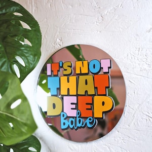 Puede incluir: Espejo redondo con la frase "IT'S NOT THAT DEEP Babe" en letras coloridas y audaces. El espejo tiene un tinte rosado y está montado en una pared blanca texturizada. Una planta verde con hojas grandes está en primer plano.