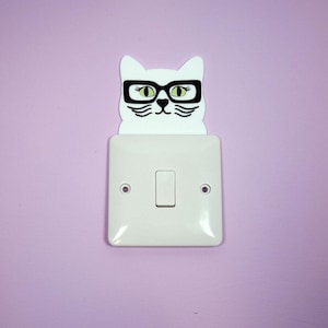 Puede incluir: Una cubierta de interruptor de luz con forma de gato blanco con gafas de montura negra y ojos verdes, montada sobre un interruptor de luz blanco en una pared lila. La cara del gato está detallada con bigotes y cejas negras.