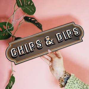 以下が含まれることがあります： 大胆なレトロフォントで「CHIPS & DIPS」と書かれた長方形の看板。看板は濃い茶色の背景に白、オレンジ、黒のアクセントがあります。大きな葉を持つ緑の植物が背景にあります。