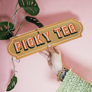 Può includere: Un'insegna rettangolare dorata con la scritta "PICKY TEA" in lettere arancioni e bianche. L'insegna ha un bordo decorativo. L'insegna è tenuta su uno sfondo rosa con piante verdi.