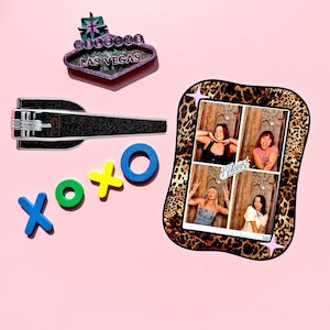 May include: A selection of decorative items on a pink backdrop. Features a Las Vegas sign magnet, a black bottle opener, XOXO magnets, and a leopard print photo frame with four photos.