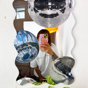 May include: A wavy-edged mirror with a silver frame reflects three disco balls, one of which is partially obscured by a person holding a phone with the text "WLR YXSZ" on the case. The person is wearing a white t-shirt and has tattoos on their arm.