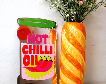 May include: A decorative mirror in the shape of a chili oil jar with the words "HOT CHILLI OIL" and a graphic of red chili peppers. A tall, orange and yellow striped vase holds a bouquet of white and pink flowers.