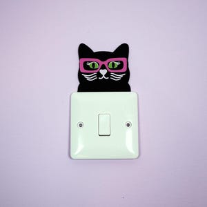 May include: A black cat light switch cover with pink glasses and green eyes, positioned above a white light switch on a lilac wall. The cat's face features white whiskers and eyebrows.