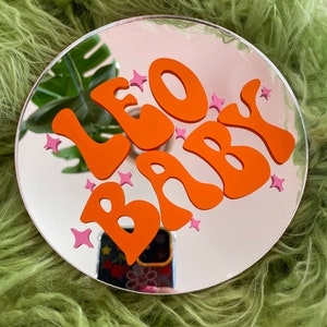 May include: A round mirror with a white background and an orange "LEO BABY" inscription in a retro font. Pink stars are scattered around the lettering.