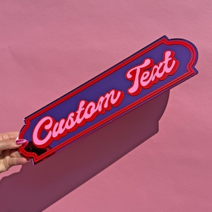 May include: A rectangular purple acrylic sign with rounded corners and a decorative border. The sign features the words Custom Text in a layered, cursive font with light pink lettering outlined in a bold red shadow against the purple surface.