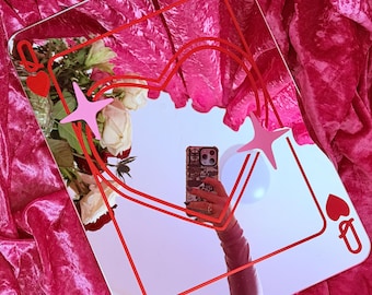 May include: A square mirror with a red outline of a playing card, featuring a heart and the Queen symbol. The mirror reflects a person holding a phone. Pink star and heart accents are included.