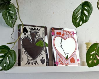 Puede incluir: Dos espejos decorativos con forma de cartas de juego. Uno presenta una forma de pica negra, el otro un corazón rosa. Los espejos se exhiben en una estantería blanca, con plantas verdes frondosas en el fondo. Los espejos miden unos 25 cm de alto.