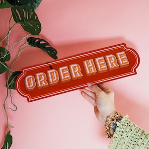 Op de afbeelding: Een rechthoekig, oranje bord met de tekst "ORDER HERE" in het wit, omlijnd in oranje. Het bord wordt tegen een roze achtergrond gehouden. Groene, bladerrijke wijnstokken bevinden zich in de linkerbovenhoek. Decoratieve rand.