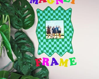 May include: A green and blue magnet frame with a photo of a group of people. The word "MAGNET" is above the frame in colorful letters, and the word "FRAME" is below. A leafy green plant is in the foreground.